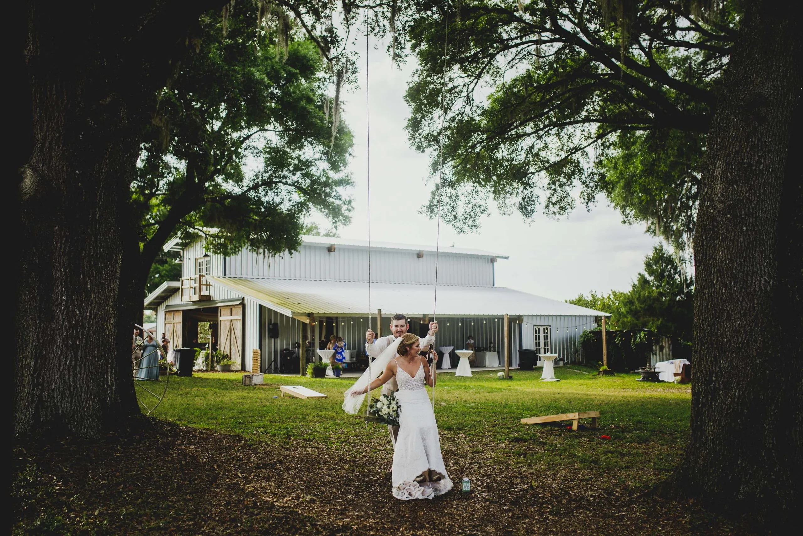 Bride on a rope swing between two live oaks while the groom pushes, with the white barn behind them