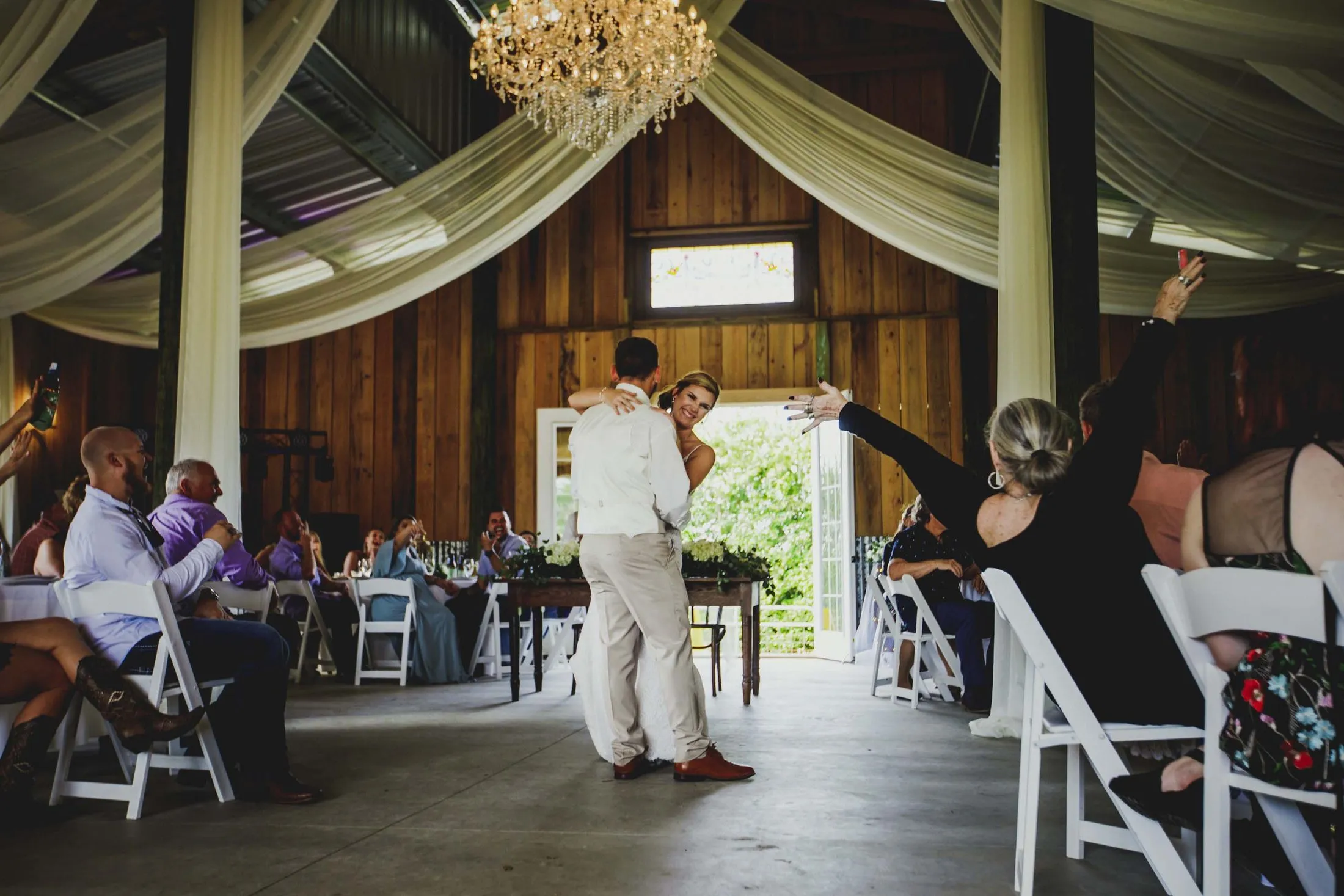 A couple’s first dance in the barn beneath the antique crystal chandelier, guests at their tables