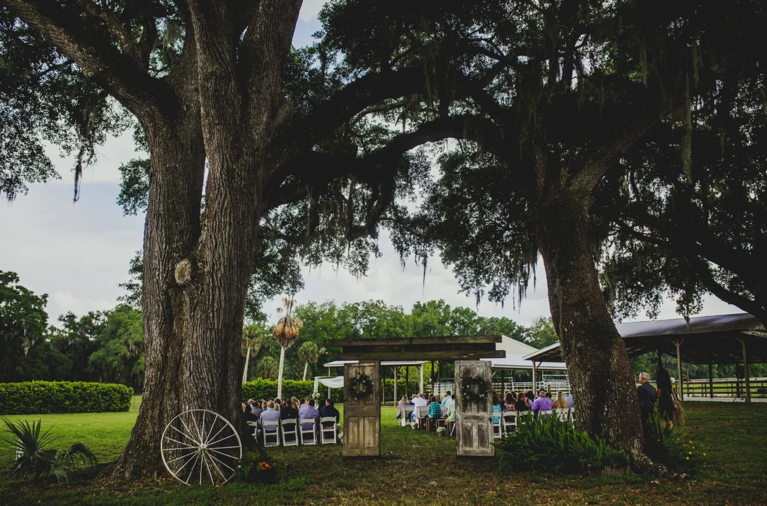 Ceremony set between two old oaks with rustic wooden doors and a wagon wheel